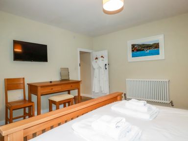 A bedroom with a wooden bed and desk with chair and stool a wall mounted tv and two white robes hanging on a door at Tretoil Farm House in Bodmin
