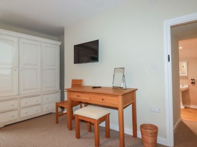 A room with a wooden desk, chair, a small cushioned stool, a wall-mounted television, a large white wardrobe, and an open doorway showing part of a bathroom at Tretoil Farm House in Bodmin