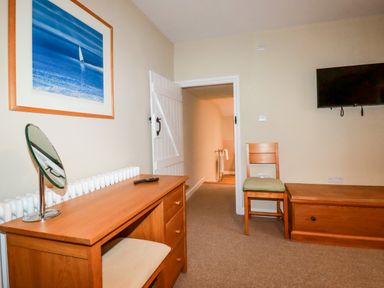 A room with a wooden desk with a mirror and remote on it a wooden chair with a green cushion a wall-mounted television a framed painting a white door and a radiator at Tretoil Farm House in Bodmin