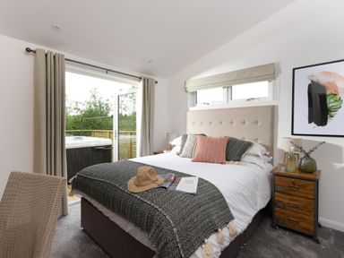 A bedroom with a bed with pillows and a blanket a bedside table with a lamp and decorations and an open door to a balcony with a hot tub at 5 Southern Place in Dobwalls