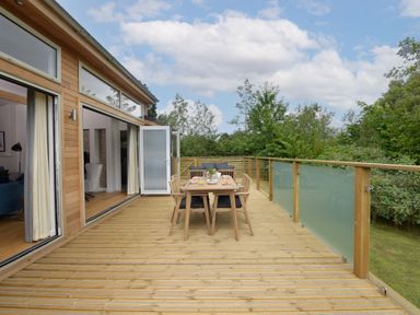 An outdoor wooden deck with a dining table and chairs next to a house with large windows and glass railing at 5 Southern Place in Dobwalls