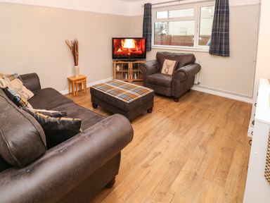 A living room with brown sofas a plaid ottoman a TV showing a fireplace and windows with curtains at Swn Y Mor in Burry Port
