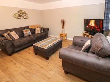 A living room with two sofas a plaid ottoman a television showing a fireplace and wooden flooring at Swn Y Mor in Burry Port