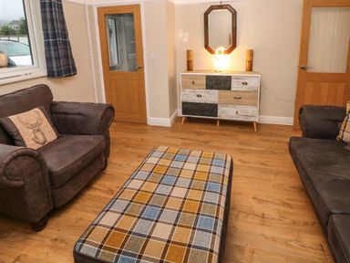 A living room with a sofa, armchair, plaid ottoman, wooden dresser with a mirror and table lamp at Swn Y Mor in Burry Port