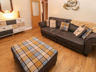 A living room with a dark sofa with plaid and deer print pillows an ottoman with plaid upholstery a wooden floor a sideboard a mirror and a door at Swn Y Mor in Burry Port