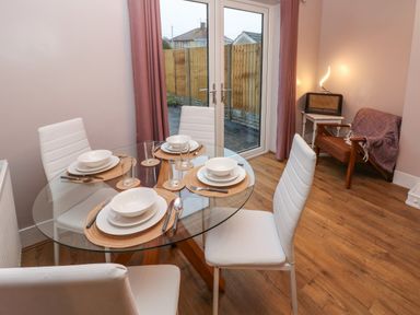 A dining room with a round glass table set for four with white chairs and a brown armchair with a pink throw at Swn Y Mor in Burry Port