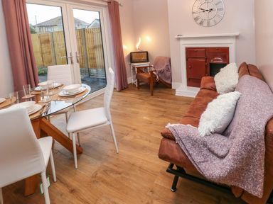 A dining area with a glass table and white chairs next to a brown sofa with cushions and a fireplace at Swn Y Mor in Burry Port