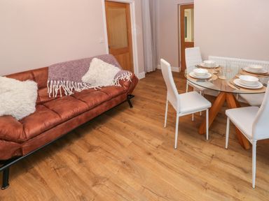A room with a brown sofa with white pillows and a purple throw next to a round glass dining table with four white chairs at Swn Y Mor in Burry Port