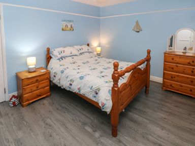 A bedroom with a wooden double bed with patterned bedding two wooden nightstands with lamps a wooden dresser with a mirror and blue walls at Swn Y Mor in Burry Port