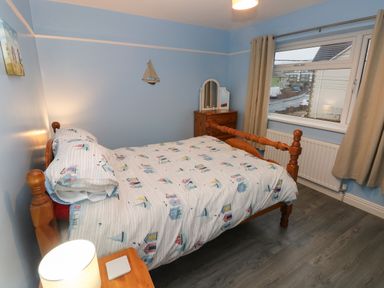 A bedroom with a wooden bed with sailboat bedding a wooden dresser with a mirror a window with curtains and a nightstand with a lamp at Swn Y Mor in Burry Port