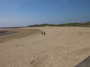 A sandy beach with three people walking and grassy dunes in the background at Swn Y Mor in Burry Port
