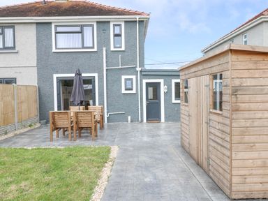 A backyard with a wooden shed wooden outdoor table and chairs with an umbrella a lawn and a grey house in Swn Y Mor in Burry Port