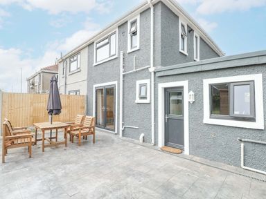 An outdoor patio area with a wooden table and chairs and an umbrella next to a two-story grey house with multiple windows and a door at Swn Y Mor in Burry Port