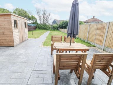 A backyard with wooden shed a patio area with wooden table and chairs and a closed umbrella at Swn Y Mor in Burry Port