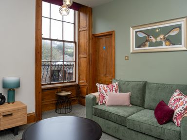 A living room with a green sofa with pillows a round black table a wooden side table with lamp and a large window at Watermouth Castle Bassett Apartment Berrynarbor near Ilfracombe