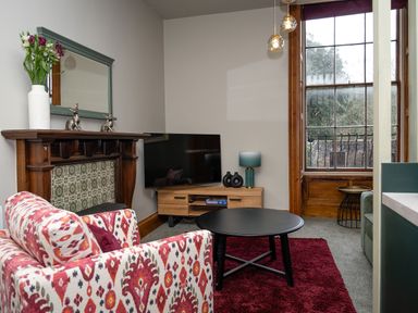 A living room with a patterned armchair a black round coffee table a wooden television stand with a lamp and decorative items a fireplace with a mirror and flowers on the mantel and large window at Watermouth Castle Bassett Apartment Berrynarbor near Ilfracombe