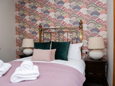 A bedroom with a brass bed frame, patterned wallpaper, two bedside tables with lamps, and folded towels on a bed with pillows and a pink quilt at Watermouth Castle Bassett Apartment Berrynarbor near Ilfracombe