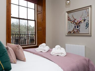 A bedroom with a bed covered with a mauve quilt and white towels near a window and deer painting on the wall at Watermouth Castle Bassett Apartment Berrynarbor near Ilfracombe