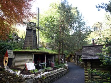 A miniature windmill and small wooden structures along a paved path in a wooded area at Watermouth Castle, Bassett Apartment in Berrynarbor near Ilfracombe