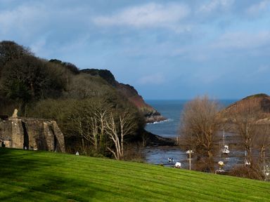 A landscape with a grassy hillside trees stone structure boats in water and hills by the sea at Watermouth Castle Rhododendron Apartment Berrynarbor near Ilfracombe