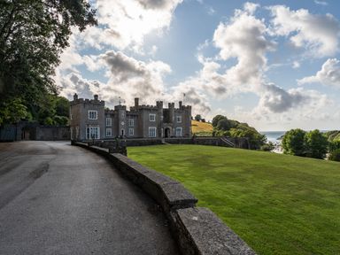 A castle with a large lawn and a driveway surrounded by trees near the sea at Watermouth Castle Rhododendron Apartment Berrynarbor near Ilfracombe
