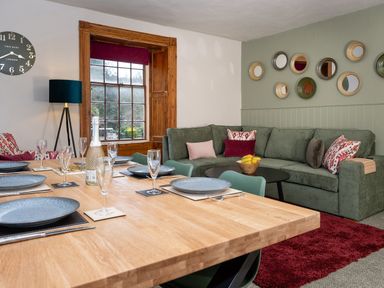 A dining area with a wooden table set with plates and glasses next to a green sofa with cushions and a round table at Watermouth Castle Rhododendron Apartment Berrynarbor near Ilfracombe
