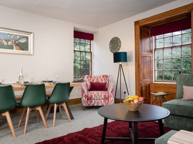 A dining area with green chairs and a wooden table alongside a sitting area with a patterned armchair a round black coffee table and large windows at Watermouth Castle Rhododendron Apartment Berrynarbor near Ilfracombe