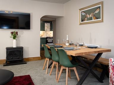 A dining room with a wooden table set with plates and glasses green chairs and a bench a television mounted on the wall and a painting of two rabbits at Watermouth Castle Rhododendron Apartment Berrynarbor near Ilfracombe