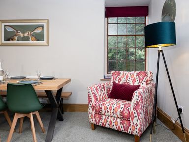A dining area with a wooden table and green chairs next to a patterned armchair with a red cushion and a floor lamp at Watermouth Castle Rhododendron Apartment Berrynarbor near Ilfracombe