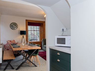 A dining area with table set for four, bench and chairs near a window with wooden frame and adjacent living room visible at Watermouth Castle Rhododendron Apartment Berrynarbor near Ilfracombe