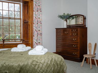 A bedroom with a bed covered by an olive green quilt and rolled white towels a wooden dresser with a mirror and a vase with green plant beside a wooden chair with two backrests at Watermouth Castle Rhododendron Apartment in Berrynarbor near Ilfracombe