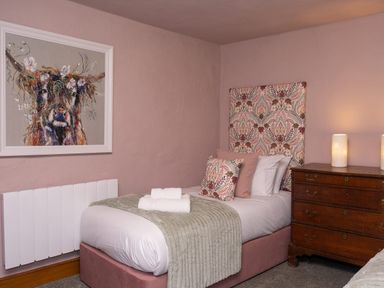 A bedroom with a single bed with patterned headboard and cushions beside a wooden chest of drawers with two lamps at Watermouth Castle Rhododendron Apartment Berrynarbor near Ilfracombe