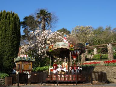 A small vintage carousel surrounded by a wooden fence with trees and flowers in the background at Watermouth Castle Rhododendron Apartment Berrynarbor near Ilfracombe
