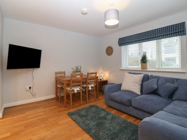 A living room with blue sofa and white cushions a wooden dining table with four chairs a wall mounted television a window with blinds and a potted plant at Ger Y Lli in Rhosneigr