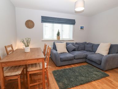 A living room with a wooden dining table and chairs a blue sectional sofa a green rug and a window with striped blinds at Ger Y Lli in Rhosneigr