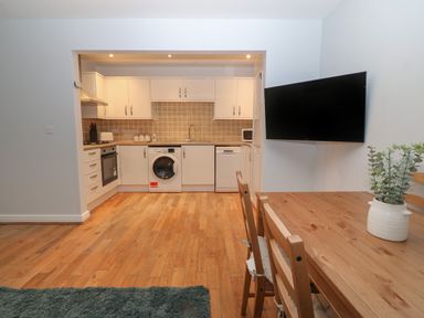 A kitchen area with white cabinets washing machine and dishwasher a wooden dining table with chairs and a wall mounted television at Ger Y Lli in Rhosneigr