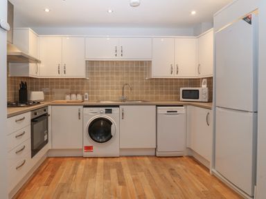 A kitchen with white cabinets a washing machine a dishwasher a microwave a refrigerator and wooden flooring at Ger Y Lli in Rhosneigr