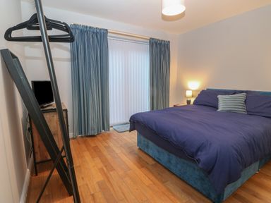 A bedroom with a bed covered in dark blue bedding a nightstand with a lamp a television on a wooden cabinet blue curtains and wooden floor at Ger Y Lli in Rhosneigr