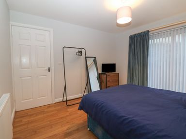 A bedroom with a bed covered with a dark blue blanket a wooden floor a door a clothing rack a tall mirror a small wooden chest of drawers with a TV on top and a window with vertical blinds and dark curtains at Ger Y Lli in Rhosneigr