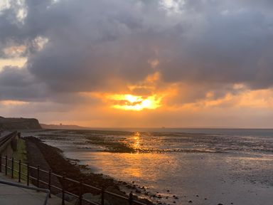 A coastal walkway with railings and rocks by the sea at sunset with clouds and the sun reflecting on the water at The Artist's Cabin By The Sea in Herne Bay