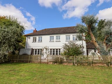 A two story house with white walls and multiple windows behind a wooden fence and trees at Culver Court Cottage in Venny Tedburn near Crediton