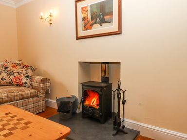 A wood burning stove with fire burning a plaid sofa with floral cushions and a wooden table at Culver Court Cottage in Venny Tedburn near Crediton