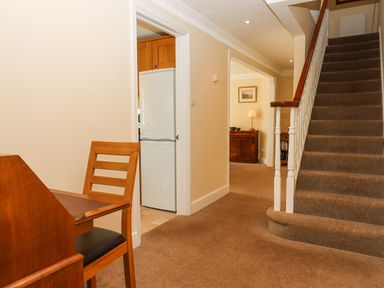 A hallway with a staircase and wooden chair next to a small desk near a kitchen with a white refrigerator at Culver Court Cottage in Venny Tedburn near Crediton