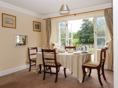 A dining room with a round table set for four near open windows and a garden at Culver Court Cottage in Venny Tedburn near Crediton