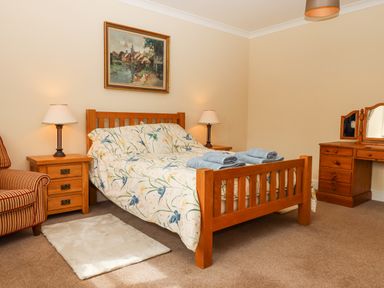 A bedroom with a wooden bed and bedside tables with lamps next to a dressing table and a striped armchair at Culver Court Cottage in Venny Tedburn near Crediton