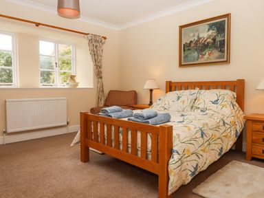 A bedroom with a wooden bed and floral bedding with two folded towels a striped armchair two nightstands with lamps a painting on the wall and a window with floral curtains at Culver Court Cottage in Venny Tedburn near Crediton