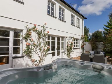 A hot tub in a garden with climbing rose plants on a white house wall and outdoor seating at Culver Court Cottage in Venny Tedburn near Crediton