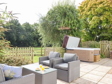 A garden patio with gray wicker chairs a glass table with food and drinks and a hot tub surrounded by wooden fencing and trees at Culver Court Cottage in Venny Tedburn near Crediton