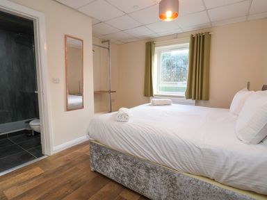 A bedroom with a double bed with white linens a window with green curtains a wall mirror an open doorway showing a bathroom at The Bevan in Bontnewydd Gwynedd