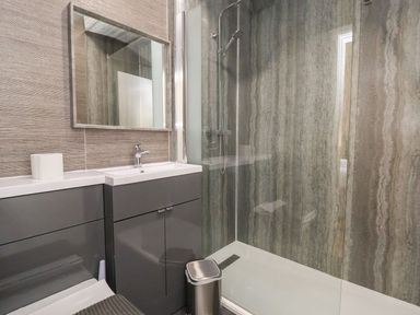 A bathroom with a gray vanity unit a mirror a roll of toilet paper a trash bin and a glass shower enclosure at The Bevan in Bontnewydd Gwynedd